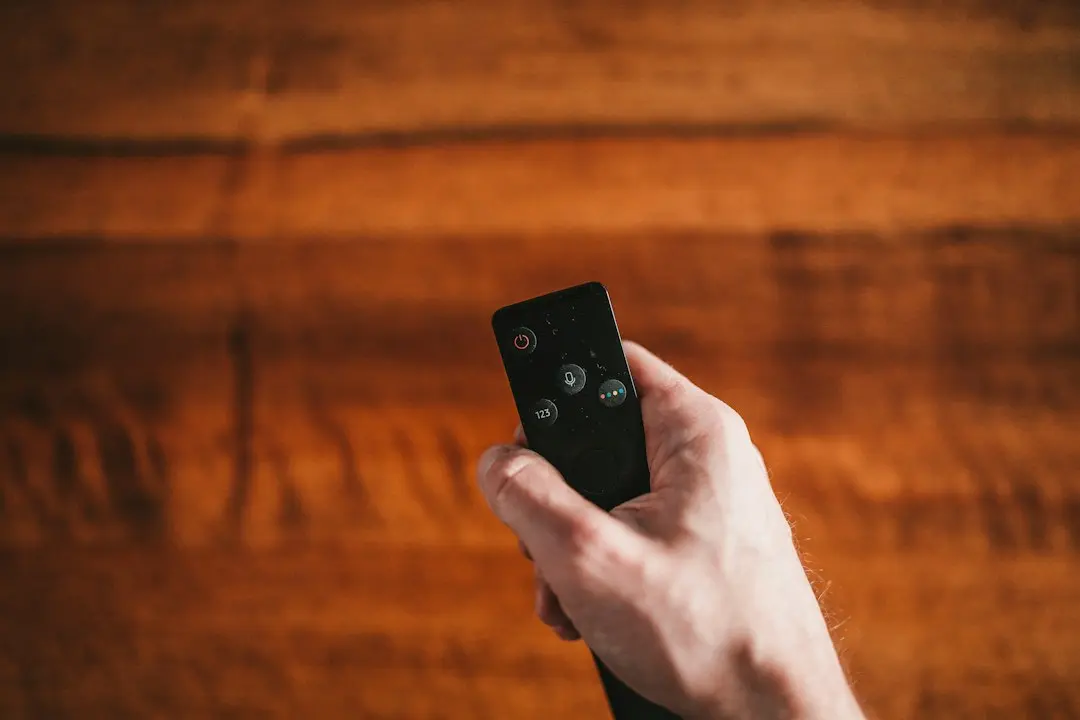 Hand holding a small black remote control against a warm wooden background